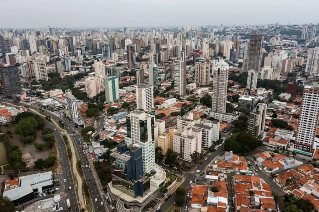 Imagem da vista aérea de Campinas mostra prédios altos e modernos e ruas com casas menores. Uma estrada larga com tráfego corre ao longo do lado esquerdo, cercada por vegetação e estruturas urbanas.