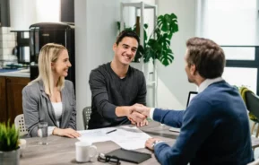 Imagem de dois homens apertando as mãos em uma mesa coberta de papéis, enquanto uma mulher está sentada perto e sorrindo para ilustrar matéria sobre como amortizar financiamento Caixa.