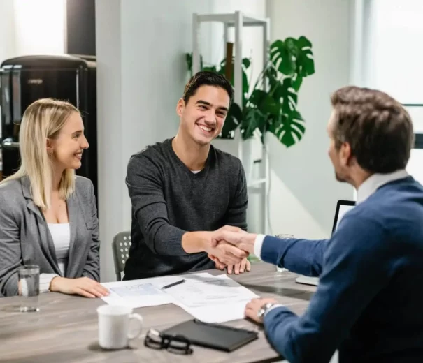 Imagem de dois homens apertando as mãos em uma mesa coberta de papéis, enquanto uma mulher está sentada perto e sorrindo para ilustrar matéria sobre como amortizar financiamento Caixa.