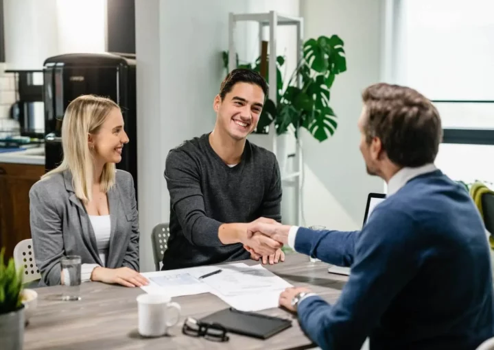 Imagem de dois homens apertando as mãos em uma mesa coberta de papéis, enquanto uma mulher está sentada perto e sorrindo para ilustrar matéria sobre como amortizar financiamento Caixa.