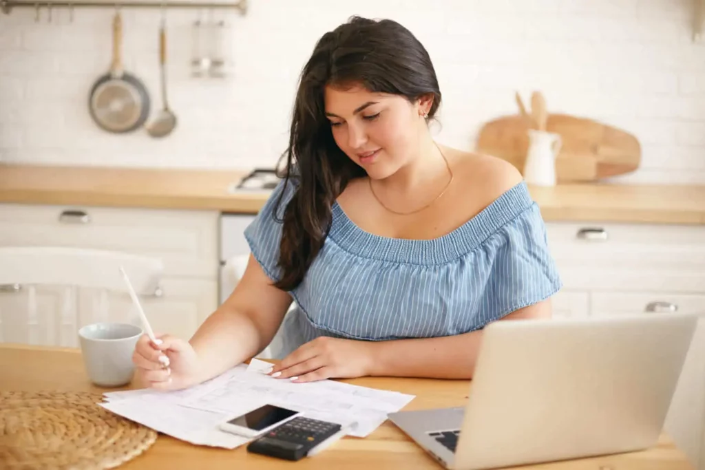 Imagem de uma mulher sentada em uma mesa de cozinha com papéis, uma calculadora e um laptop, escrevendo com uma caneta. Ela parece concentrada, com uma caneca ao seu lado e utensílios de cozinha visíveis ao fundo.