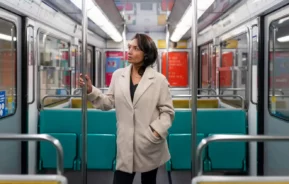 Imagem de uma mulher com um casaco bege dentro de um vagão de metrô vazio, olhando para os lados com uma expressão pensativa. Os assentos são azul-turquesa, e o ambiente parece limpo e moderno.