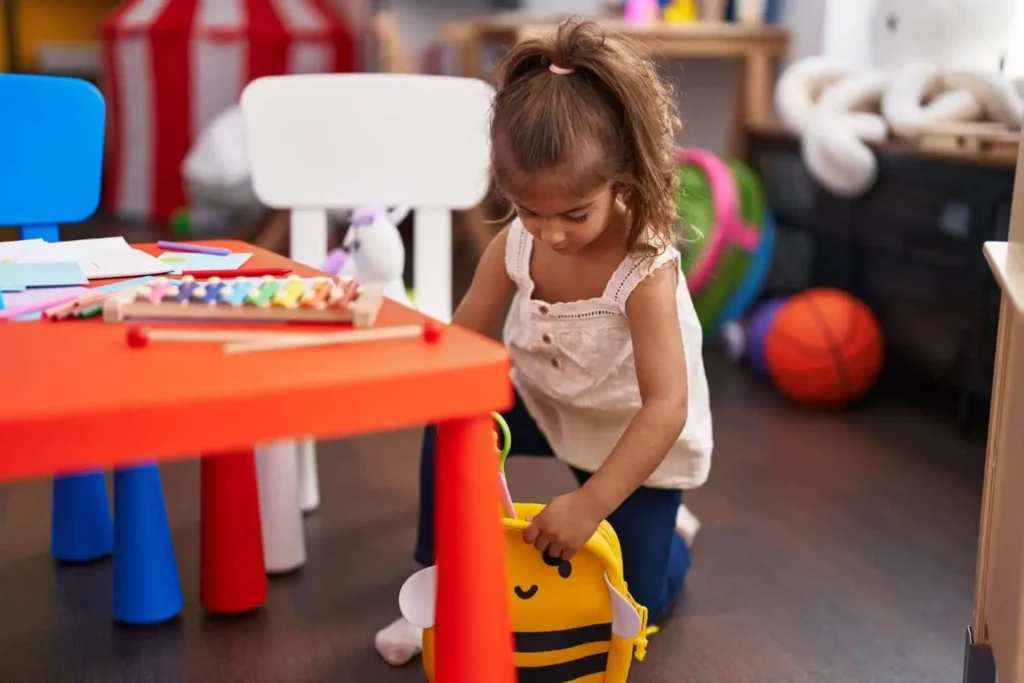 Imagem de uma menina em uma brinquedoteca de condomínio segurando uma mochila amarela com um desenho de abelha. Uma mesa vermelha com materiais de arte e um xilofone está próxima, e brinquedos e uma bola de basquete podem ser vistos ao fundo.