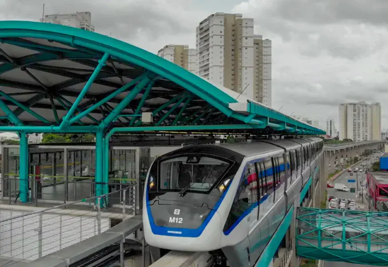 Imagem de um trem de metrô moderno da cidade de São Paulo com detalhes em azul e preto chegando a uma estação urbana elevada, com prédios de apartamentos altos e um céu nublado ao fundo.