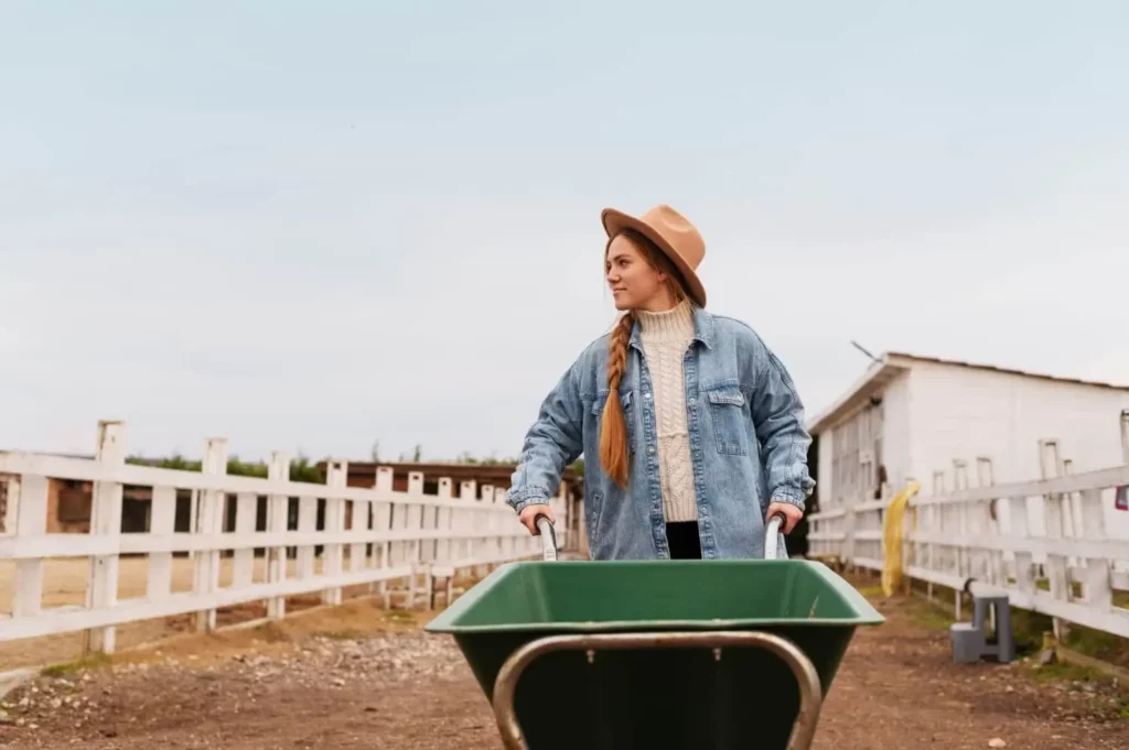 Imagem de uma mulher com jaqueta jeans e chapéu empurrando um carrinho de mão verde ao longo de um caminho de terra entre cercas brancas, com celeiros e edifícios de fazendas ao fundo.