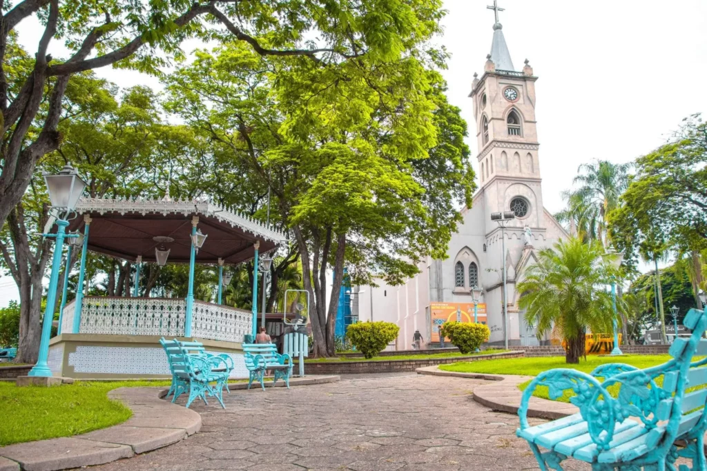 Imagem de Jaguariúna, em São Paulo, mostra uma praça arborizada com bancos turquesa, um gazebo branco e uma igreja histórica branca com uma torre de relógio ao fundo, cercada por vegetação e palmeiras em um dia ensolarado.
