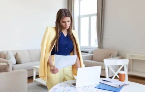 Imagem de uma mulher vestindo um terno amarelo em pé ao lado de uma mesa, olhando para os papéis em sua mão, para ilustrar matéria sobre locatário solidário. Um laptop, documentos e uma xícara de café estão sobre a mesa em uma sala de estar moderna e iluminada com grandes janelas.
