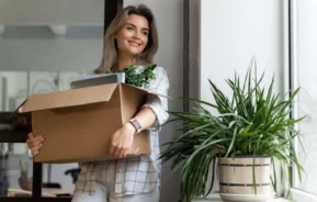 Imagem de uma mulher sorridente dentro de casa segurando uma caixa de papelão com uma planta e outros itens durante uma mudança. Ela está perto de uma janela com outro vaso de planta no parapeito.