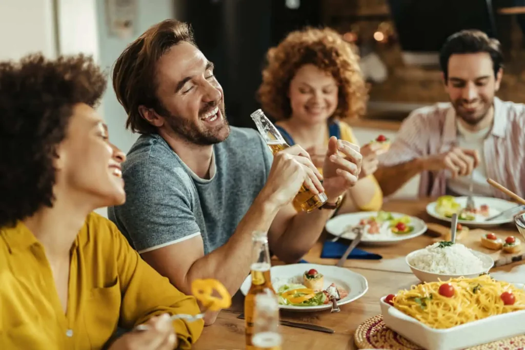 Imagem de quatro amigos sentados ao redor de uma mesa, rindo e fazendo uma refeição juntos. Uma pessoa segura uma garrafa de cerveja e há pratos de comida e macarrão sobre a mesa. A atmosfera é alegre e descontraída.