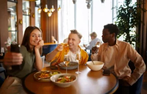 Imagem de três amigos sentados em uma mesa de restaurante, sorrindo e saboreando alimentos e bebidas, para ilustrar matéria sobre os melhores restaurantes em São Leopoldo. Uma pessoa tira uma selfie enquanto os outros posam e comem, cercados por pratos e copos em um interior moderno e iluminado.