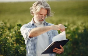 Imagem de um homem mais velho, com cabelos grisalhos e barba, usando óculos e camisa xadrez, ao ar livre em um campo verde, segurando uma prancheta e folheando papéis enquanto olha para baixo pensativamente para ilustrar matéria sobre o que é ITR.