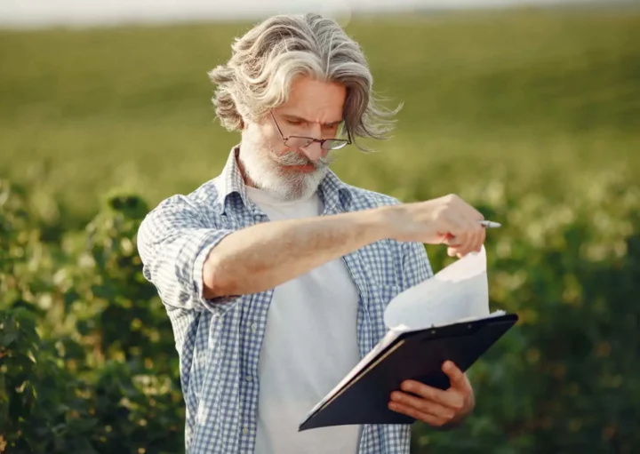 Imagem de um homem mais velho, com cabelos grisalhos e barba, usando óculos e camisa xadrez, ao ar livre em um campo verde, segurando uma prancheta e folheando papéis enquanto olha para baixo pensativamente para ilustrar matéria sobre o que é ITR.