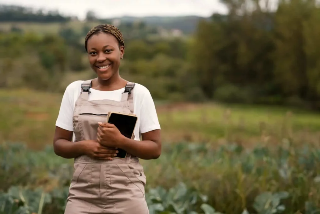 Imagem de uma jovem mulher vestindo macacão e camiseta branca está ao ar livre em um campo, sorrindo e segurando um tablet. Vegetação verde e árvores são visíveis ao fundo.