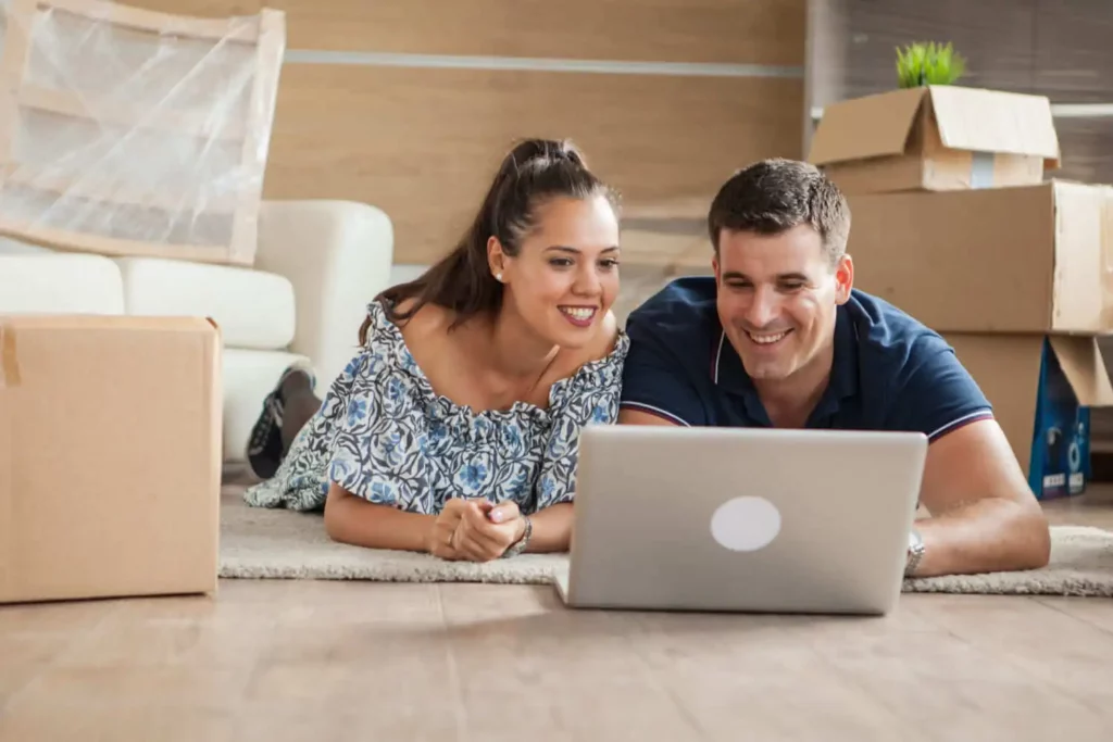 Imagem de um casal sorridente deitado no chão entre caixas de mudança, olhando para um laptop juntos em uma sala iluminada durante uma mudança.
