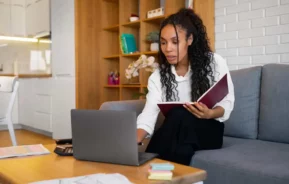 Imagem de uma mulher de cabelos cacheados sentada em um sofá cinza, segurando uma pasta e trabalhando em um laptop em uma sala de estar moderna. Papéis, notas adesivas e uma calculadora estão sobre a mesa de centro de madeira à sua frente.
