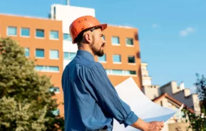 Imagem de um arquiteto usando um capacete de construção laranja e camisa azul segura plantas arquitetônicas enquanto está ao ar livre em frente a um edifício moderno para ilustrar matéria sobre o que é RRT. Árvores e um céu azul claro estão ao fundo.