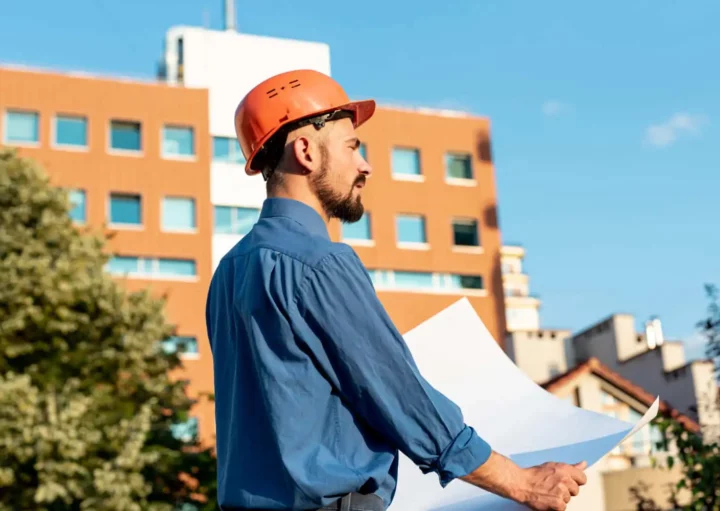 Imagem de um arquiteto usando um capacete de construção laranja e camisa azul segura plantas arquitetônicas enquanto está ao ar livre em frente a um edifício moderno para ilustrar matéria sobre o que é RRT. Árvores e um céu azul claro estão ao fundo.