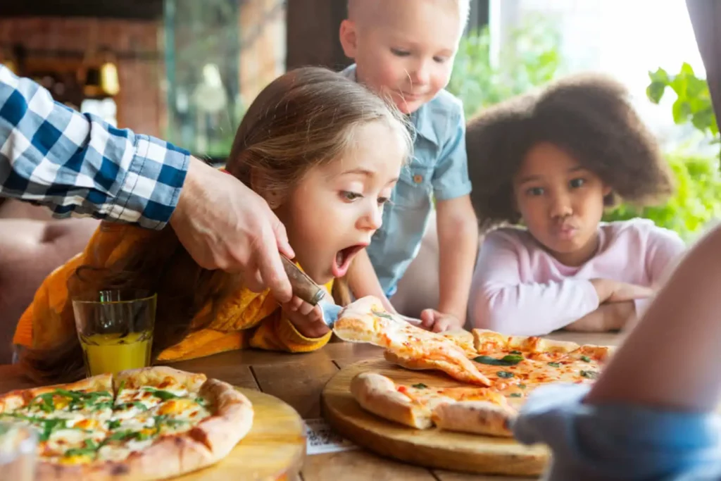 Imagem de três crianças sentadas em uma mesa com pizzas. Uma menina alcança animadamente uma fatia que está sendo servida, enquanto outra menina e um menino observam. Uma mão com um cortador de pizza serve a pizza enquanto bebidas estão em cima da mesa