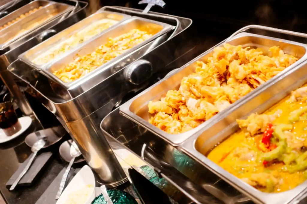 Imagem de bandejas de buffet de aço inoxidável cheias de vários pratos quentes, incluindo frituras e molhos, exibidas em um balcão de serviço com colheres próximas.