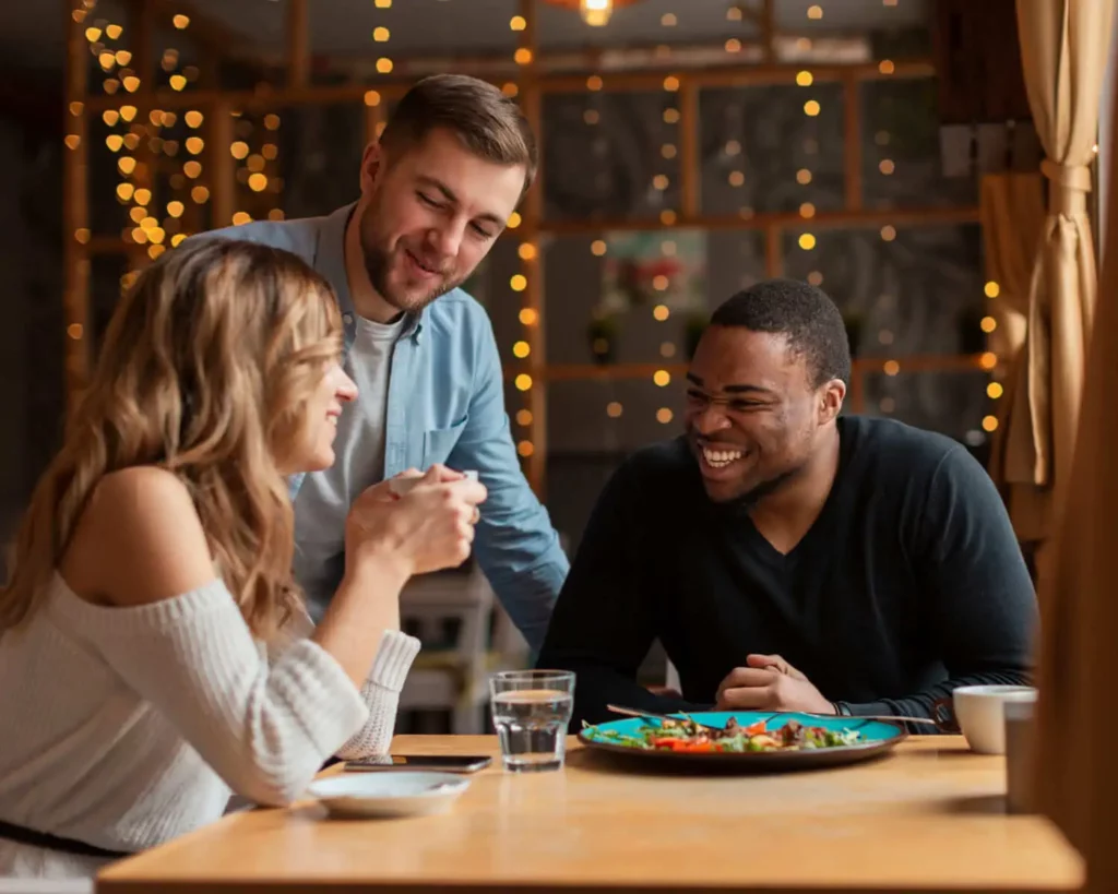 Imagem de um garçom sorridente ao lado de uma mesa onde uma mulher e um homem estão conversando e rindo alegremente, com pratos de comida e copos de água sobre a mesa, para ilustrar matéria sobre o que fazer em São Leopoldo à noite.