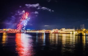 Imagem de fogos de artifício coloridos iluminando o céu noturno sobre uma cidade à beira-mar, com reflexos cintilantes na água calma e edifícios iluminados ao fundo, para ilustrar matéria sobre onde passar o Réveillon 2025