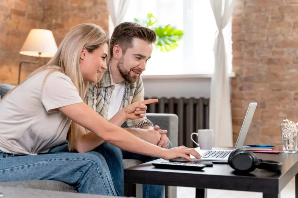Imagem de uma mulher e um homem sorridentes sentados em um sofá em uma sala de estar aconchegante, ambos olhando para um laptop sobre a mesa de centro. A mulher digita enquanto o homem aponta para a tela. Uma caneca, fones de ouvido e uma planta estão por perto.