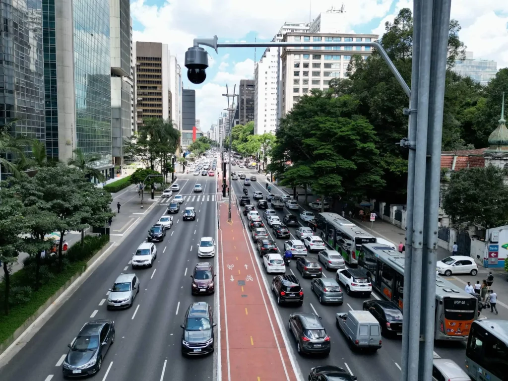 Imagem da Avenida Paulista, em São Paulo, mostra ruas movimentadas com várias faixas de carros, algumas com tráfego intenso, e uma ciclovia vermelha no centro. Prédios altos e árvores se alinham em ambos os lados e uma câmera de segurança está montada em um poste no primeiro plano.