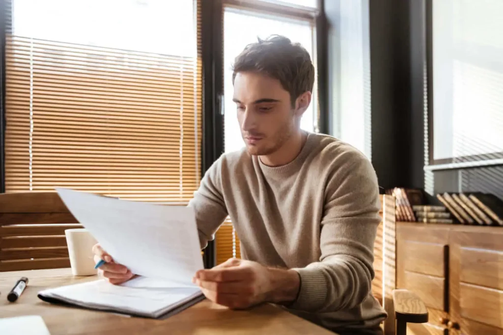 Imagem de um jovem sentado em uma mesa de madeira, lendo papéis, para ilustrar matéria sobre regularização fundiária urbana. Ele usa um suéter bege e está concentrado nos documentos. A luz do sol atravessa as persianas da janela atrás dele. Uma caneca e uma caneta estão sobre a mesa.