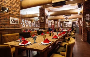 Imagem de um restaurante aconchegante com paredes de tijolos, mesas de madeira com guardanapos, copos e pratos vermelhos para ilustrar matéria sobre os restaurantes em Americana, no interior de São Paulo. O ambiente apresenta decoração vintage, fotos emolduradas nas paredes e iluminação aconchegante para criar uma atmosfera acolhedora.