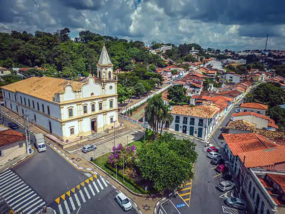 Imagem da vista aérea de Santana da Parnaíba, no interior do estado de São Paulo, mostra uma igreja branca, telhados vermelhos, ruas arborizadas, carros estacionados e vegetação sob um céu parcialmente nublado.