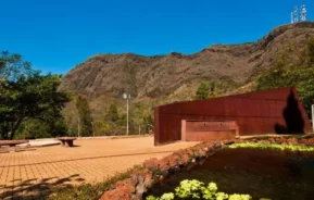 Imagem de de um edifício cor de ferrugem em uma área do parque da Serra do Curral, em BH, cercado por vegetação e colinas sob um céu azul claro.