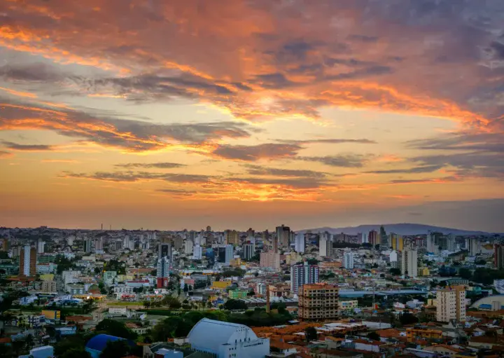 Imagem da vista aérea de Sorocaba mostra vários edifícios ao pôr do sol com nuvens em laranja e amarelo. O sol está se pondo no horizonte, lançando uma luz colorida sobre a paisagem urbana.