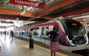 Um trem moderno chega à plataforma de uma estação movimentada com pessoas esperando. Uma placa vermelha acima diz Embarque Pirajá. A plataforma tem marcações de segurança e vários passageiros estão visíveis.