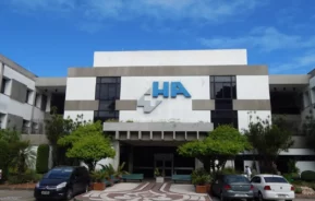 Um moderno edifício hospitalar com HA e um símbolo de cruz azul na frente. Vários carros estão estacionados na frente, com árvores verdes e um céu azul ao fundo.