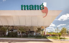 Vista externa de um restaurante com uma grande placa onde se lê mané em letras verdes e um destaque em vermelho acima da entrada, com assentos ao ar livre, palmeiras e um céu claro com nuvens.