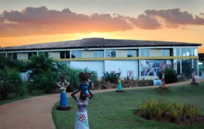 Um edifício com telhado de telhas ao pôr do sol, cercado por grama, palmeiras e esculturas coloridas de mulheres segurando vasos na cabeça ao longo de uma passarela pavimentada.