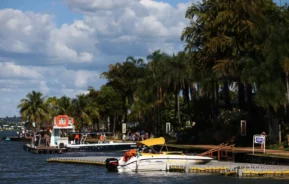 Uma lancha branca com capota amarela está atracada em uma orla com palmeiras. As pessoas caminham no píer e vários barcos são visíveis sob um céu parcialmente nublado.