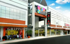 Um moderno shopping center com grandes janelas de vidro, letreiros coloridos onde se lê Nova Belas Flor, lojas de roupas e de varejo como a Grippon e um céu azul claro acima.