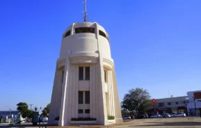 Um edifício alto, branco e cilíndrico com colunas verticais e janelas fica em uma praça sob um céu azul claro, cercado por árvores, carros e lojas próximas.