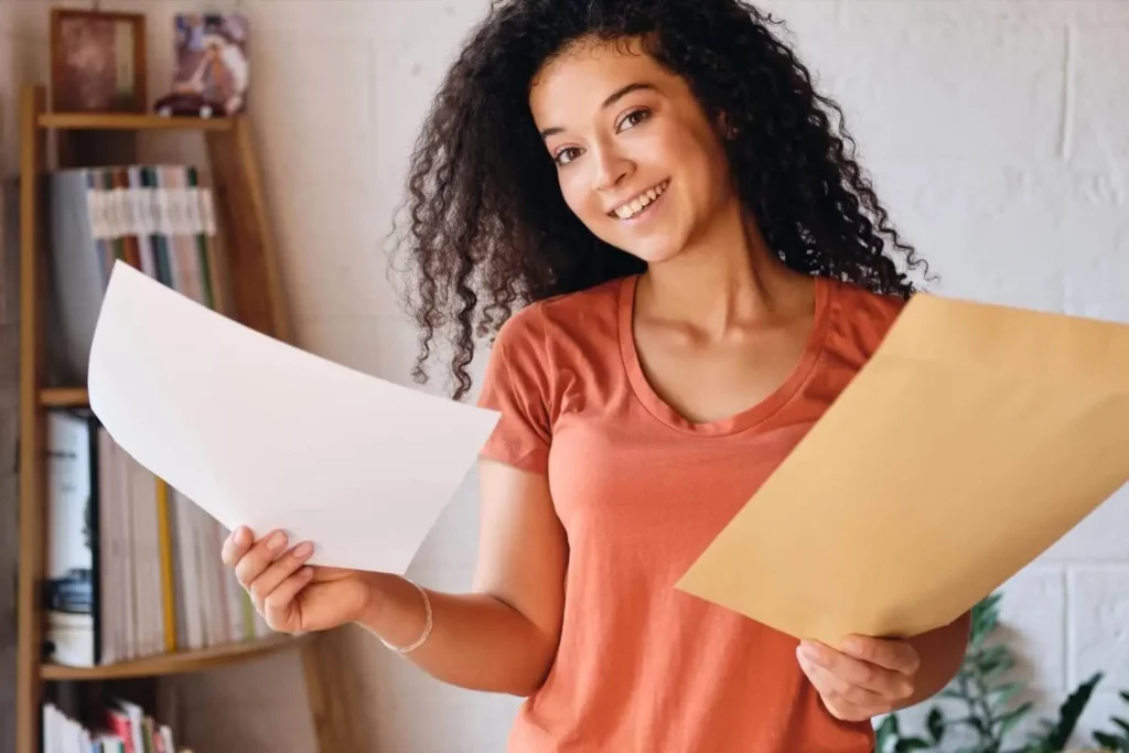 Imagem de uma mulher sorridente com cabelos cacheados segurando uma folha de papel em branco em uma das mãos e um envelope grande na outra, em pé, dentro de casa, em frente a uma estante de livros.