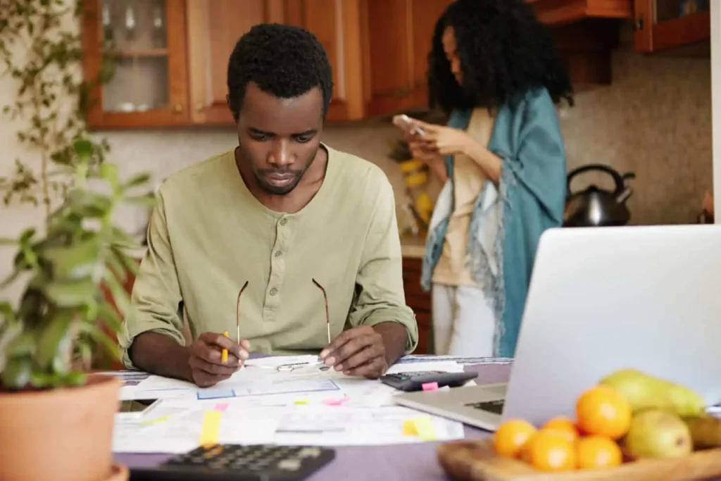 Imagem de um homem sentado em uma mesa de cozinha trabalhando com papéis, um laptop e calculadoras, enquanto uma mulher está ao fundo usando seu telefone. Frutas e um vaso de plantas também estão sobre a mesa.