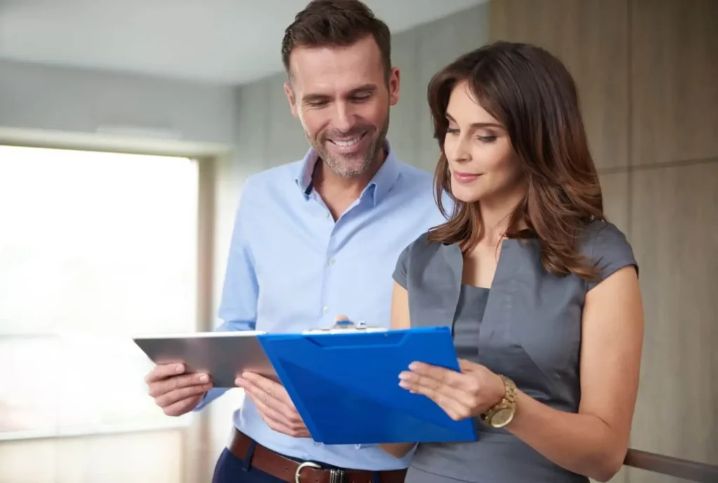 Imagem de um homem e uma mulher em trajes de negócios juntos, sorrindo e analisando documentos em pranchetas em um ambiente de escritório.