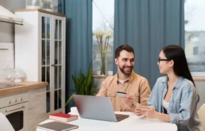 Imagem de um homem e uma mulher sentados à mesa em uma cozinha iluminada, sorrindo e conversando. Um laptop, um notebook e livros estão sobre a mesa. Grandes janelas e plantas estão ao fundo.