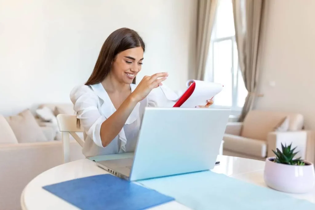 Imagem de uma mulher sentada em uma mesa, sorrindo enquanto examina papéis em uma pasta em frente a um laptop aberto. A sala é iluminada por luz natural, com um sofá, cortinas e uma planta próxima.