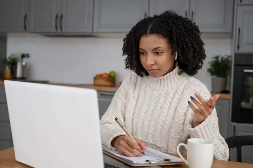 Imagem de uma mulher de suéter branco sentada em uma mesa com um laptop, escrevendo em um caderno e fazendo gestos com a mão. Ela usa fones de ouvido e tem uma caneca por perto. 