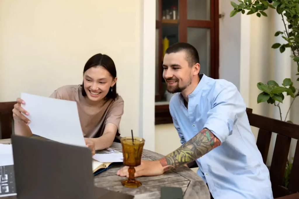 Imagem de duas pessoas sentadas em uma mesa ao ar livre, sorrindo enquanto olham alguns papéis juntas na frente de um laptop. Uma bebida com canudo e livros estão sobre a mesa, e há plantas ao fundo.