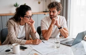 Imagem de duas pessoas sentadas em uma mesa com documentos, laptop, telefone, caneca e copo d'água, parecendo concentradas enquanto analisam documentos juntas em uma sala moderna e iluminada, para ilustrar matéria sobre quanto tempo demora a contemplação no consórcio