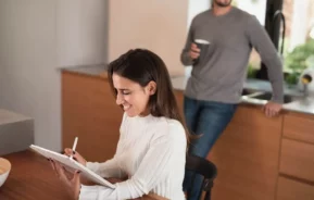 Imagem de uma mulher sentada em uma mesa, sorrindo e escrevendo em um tablet digital com uma caneta, enquanto um homem está na cozinha atrás dela segurando uma caneca. O cenário parece ser uma cozinha doméstica moderna.