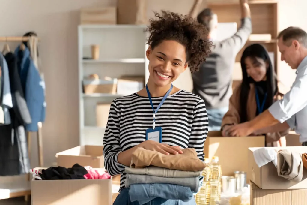 Imagem de uma mulher sorridente de cabelos cacheados, vestindo uma camisa listrada e um crachá, segura roupas dobradas. Atrás dela, pessoas empacotam caixas com alimentos e suprimentos em uma sala iluminada e organizada.