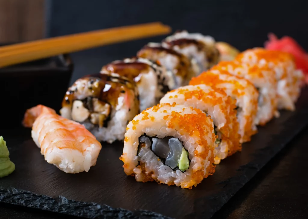 Imagem de um close-up de diversos rolos de sushi e nigiri em um prato de ardósia preta, incluindo rolos com ovas de laranja, abacate e enguia com molho, com um pedaço de nigiri de camarão, wasabi e hashis ao fundo.